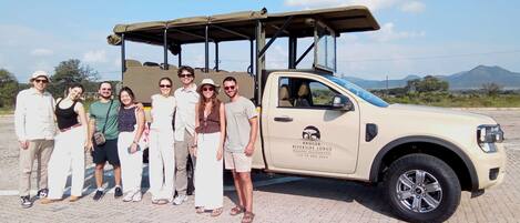 A group of people standing in front of a beige SUV with a canopy, parked on a paved area with a mountainous landscape in the background.