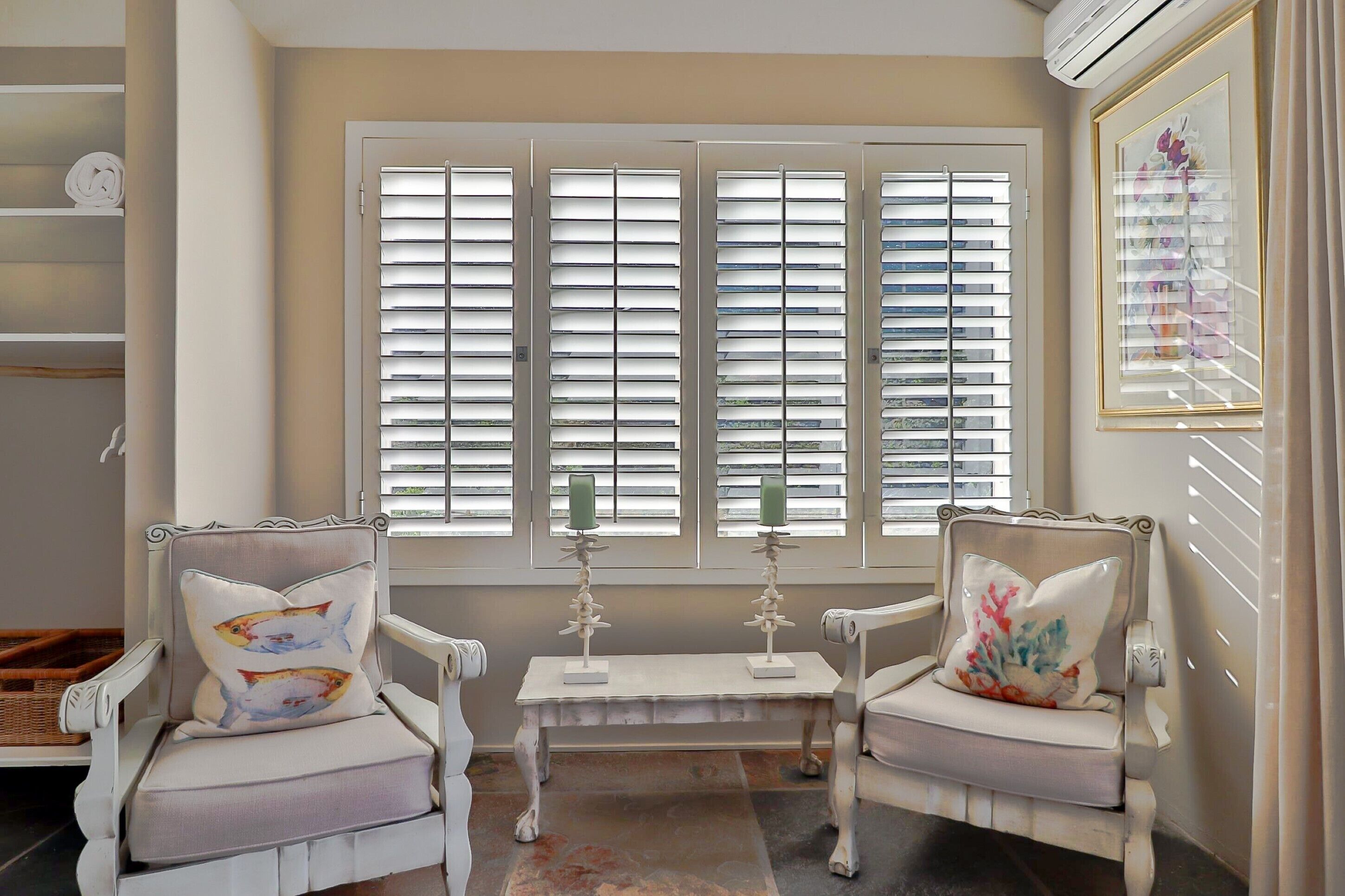A room with two white armchairs, a small table, and a bench. There are three windows with shutters, and a framed picture on the wall.