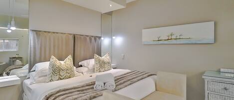 A modern hotel room with a large bed, a bedside table, a dresser, and a painting on the wall.
