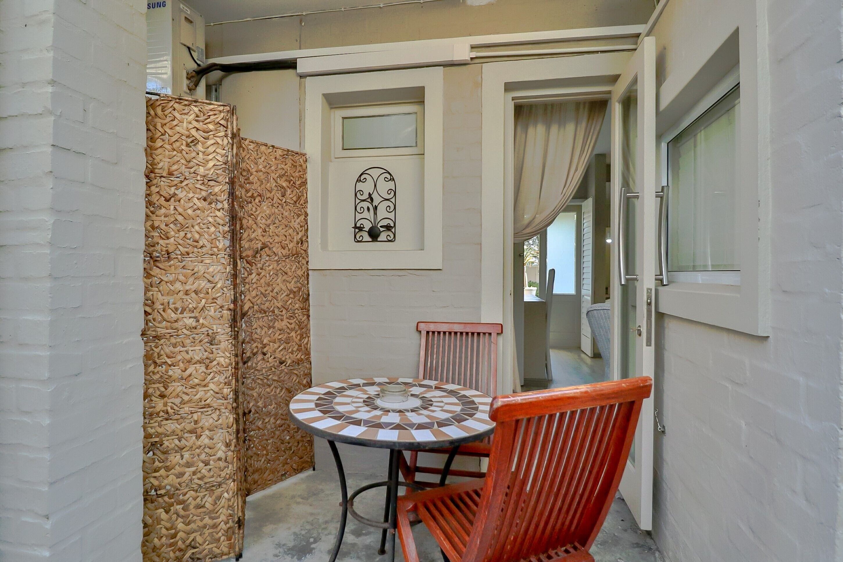 A small outdoor seating area with a round table and a single chair, a decorative wall panel, and a door leading to another room.