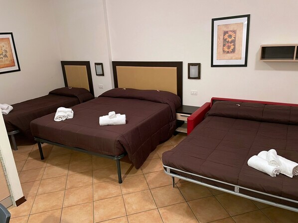 A hotel room with three beds, each with a brown bedspread and white towels. There are framed pictures on the walls and a small shelf.