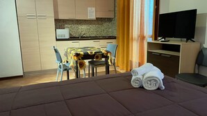 A hotel room with a bed, a TV, a desk, a chair, a table with a sunflower tablecloth, and a kitchen area. - Residence Cimone (Riolunato)