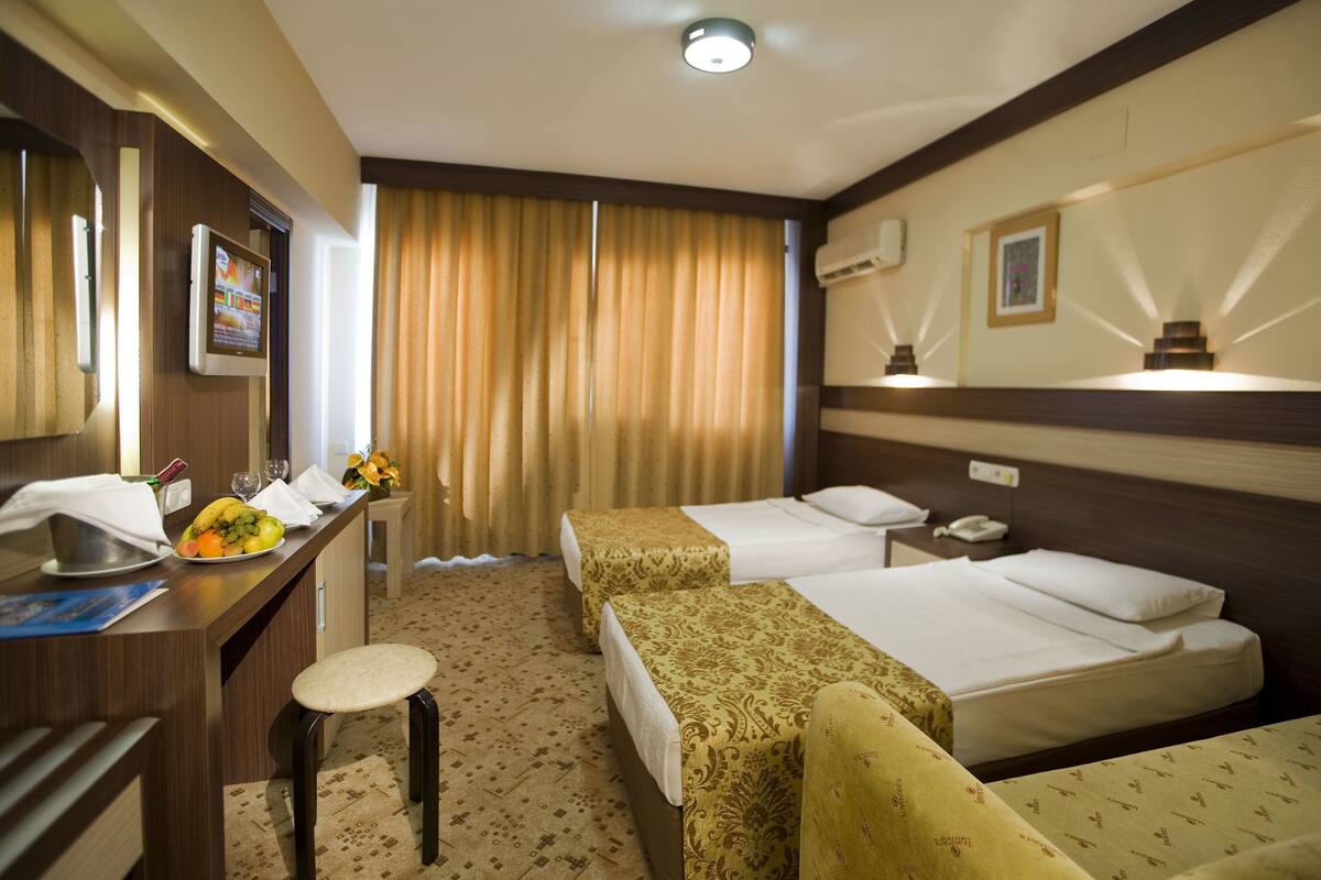 A hotel room with two beds, a desk with a fruit bowl, a mirror, and a window with curtains.