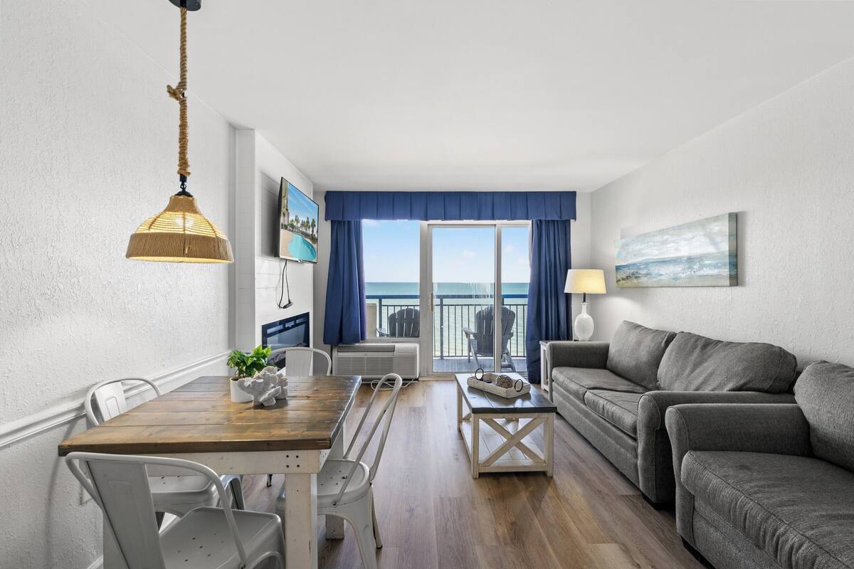 A living room with a wooden dining table, white chairs, a grey sofa, and a view of the sea through a large window.