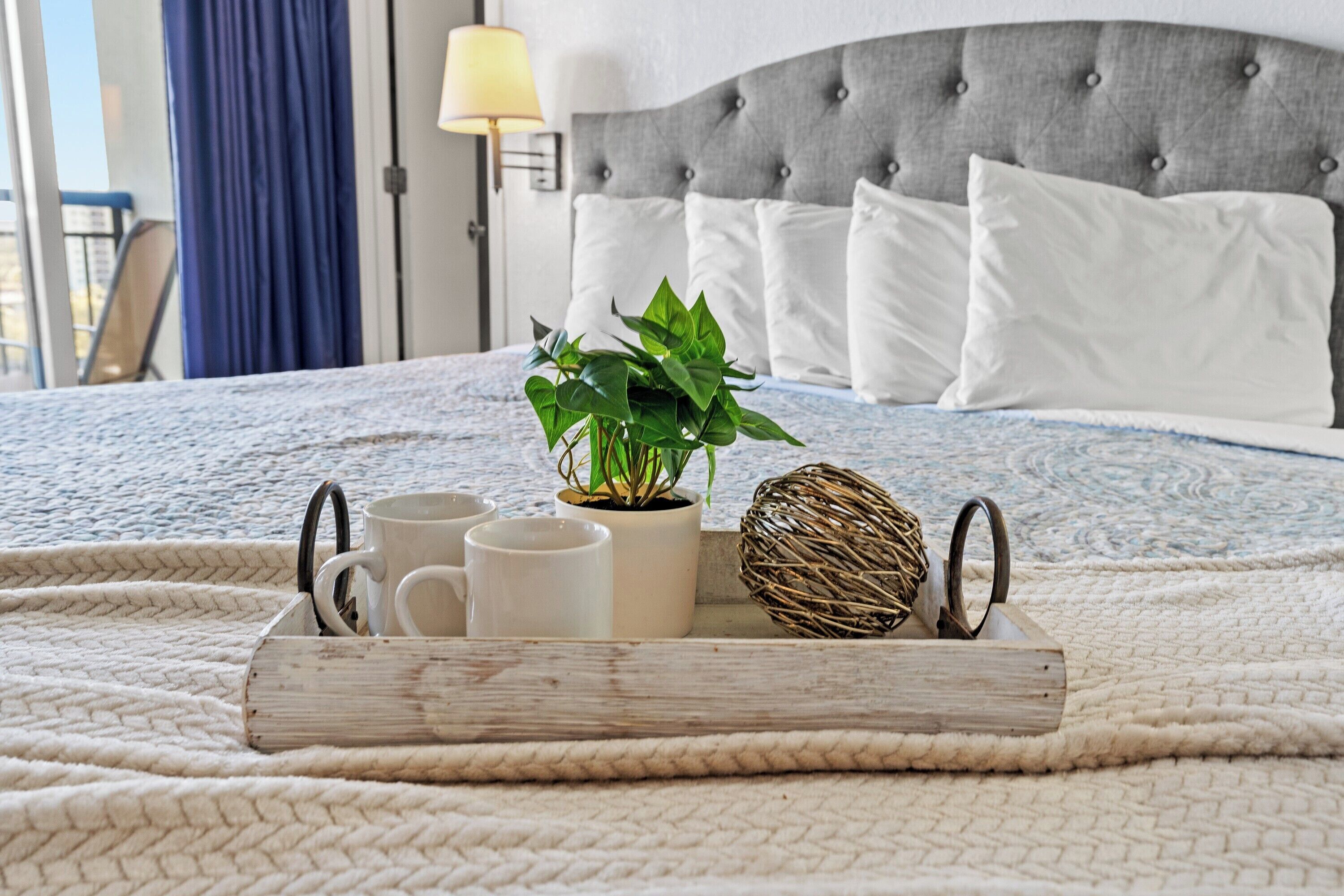 A neatly made bed with a tray of mugs, a potted plant, and a decorative item on a textured blanket.