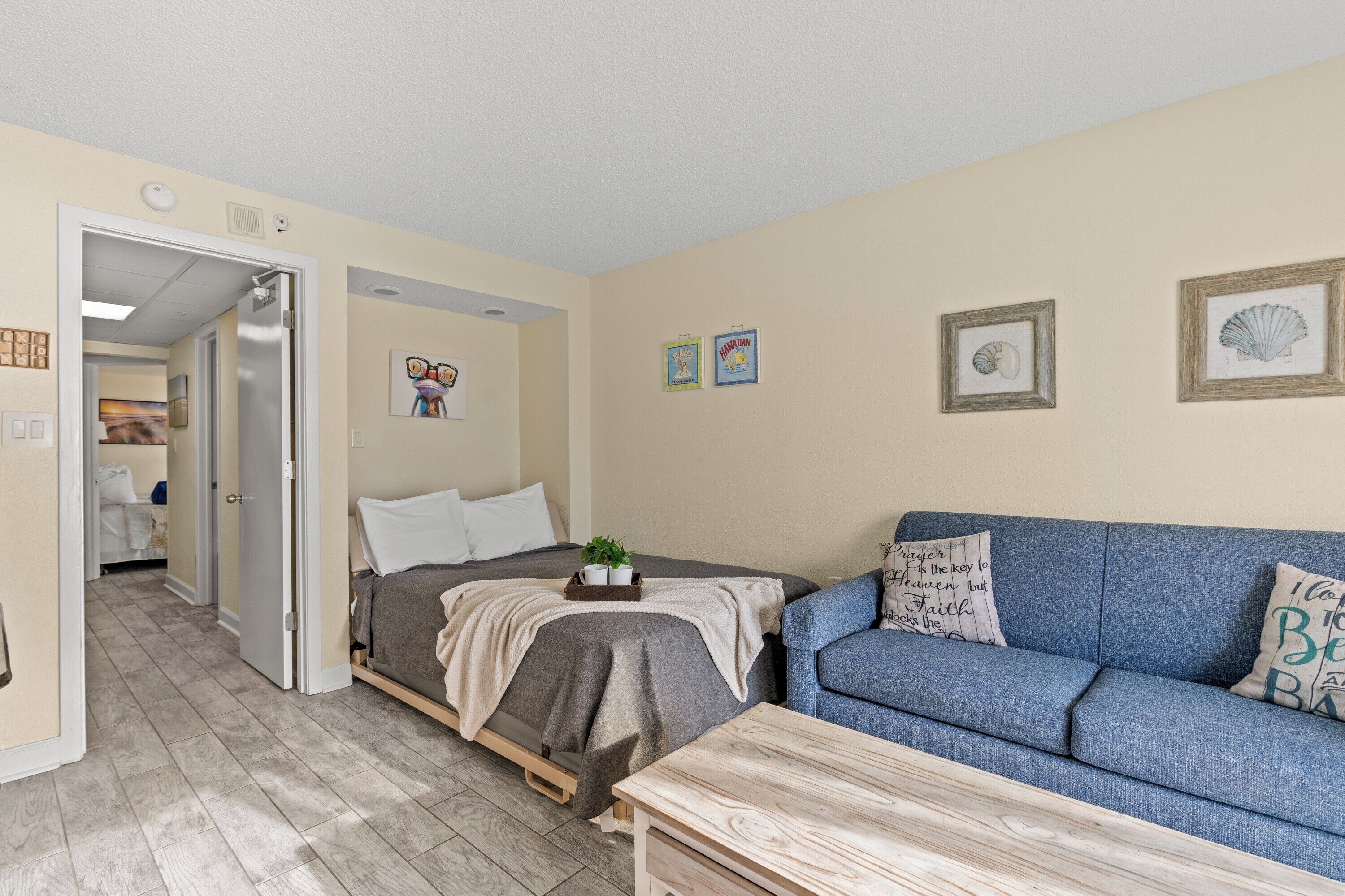 A bedroom with a bed, a blue sofa, a wooden table, and a door leading to another room.
