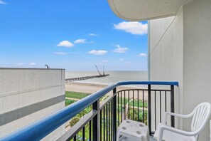 Varanda com vista para a praia e o cais, cadeiras de plástico branco e uma mesa de concreto.
