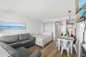Quarto moderno e bem iluminado com sofá cinza, cama, mesa de jantar com cadeiras e um quadro com uma cena de praia.