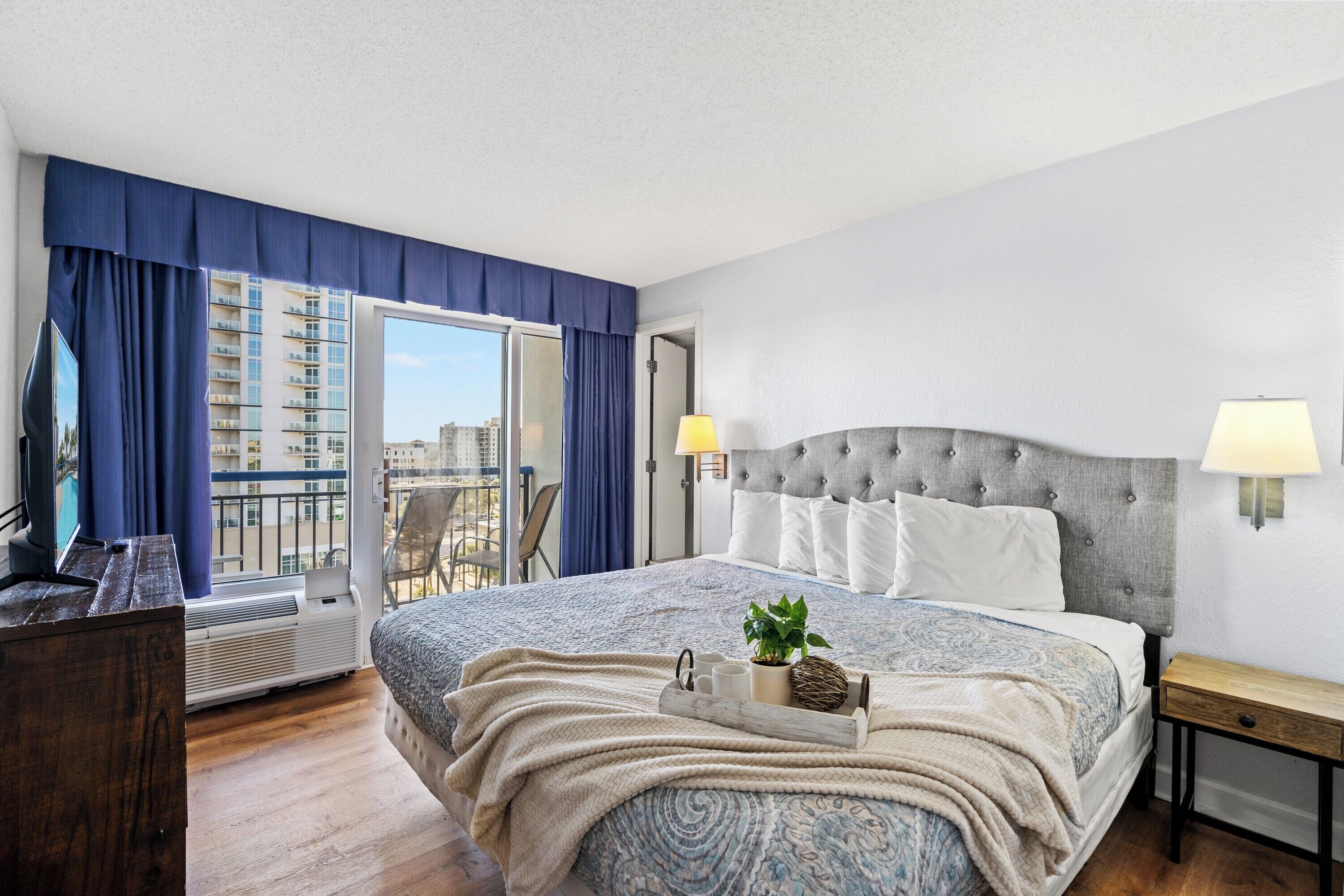 A hotel room with a large bed, a desk, a chair, a TV, and a balcony with a view of the city.