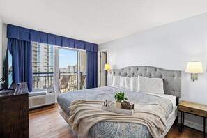 A hotel room with a large bed, a desk, a chair, a TV, and a balcony with a view of the city. - Boardwalk Beach Resort by Elliott Beach Rentals (Myrtle Beach)