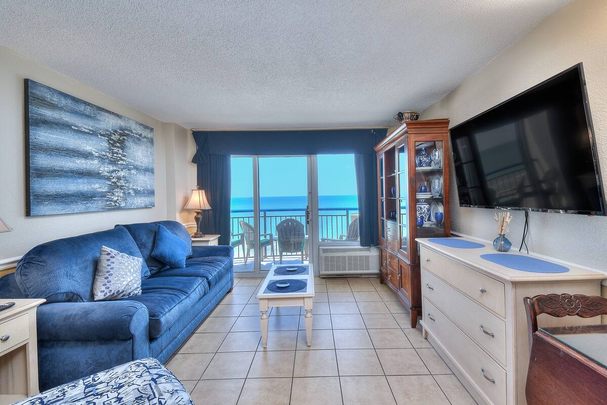 A living room with a blue sofa, a coffee table, a TV, and a view of the ocean.