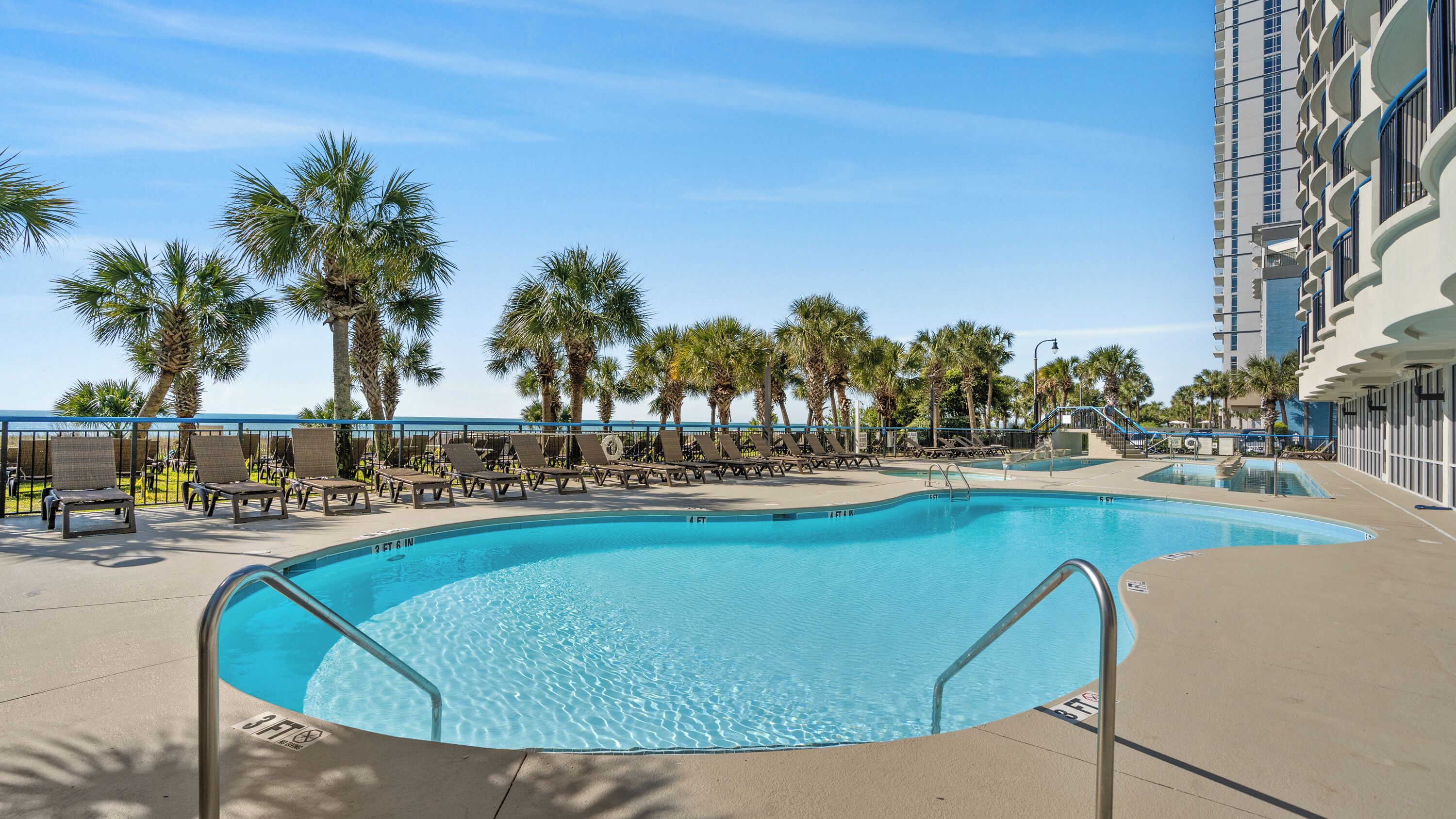 A swimming pool with lounge chairs and palm trees, surrounded by a building with balconies.