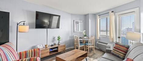 A modern living room with a sofa, armchair, coffee table, and a flat-screen TV mounted on the wall. There is a dining area with a table and chairs, and large windows with curtains.