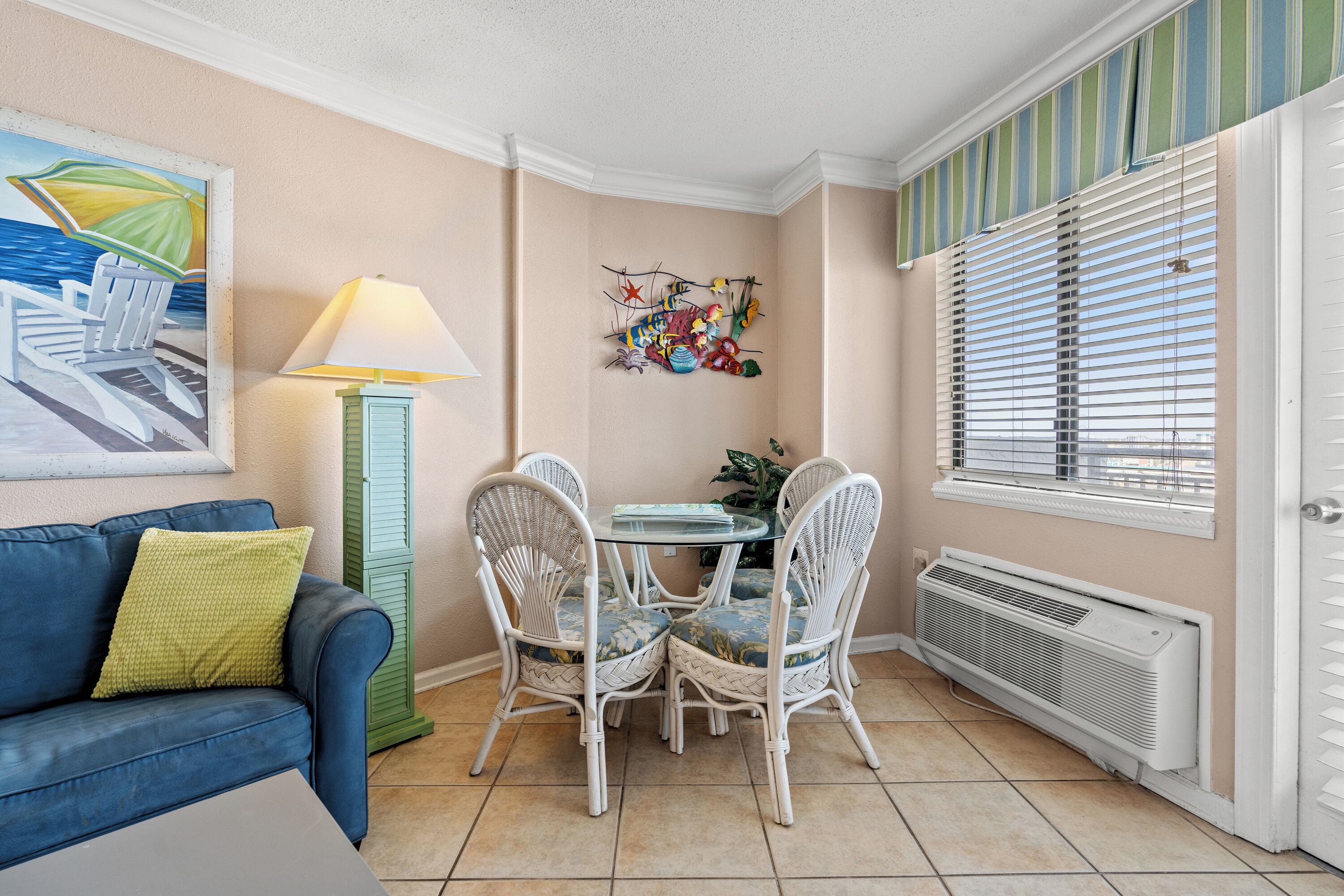 A dining area with a table and chairs, a lamp, a painting, and a window with blinds.