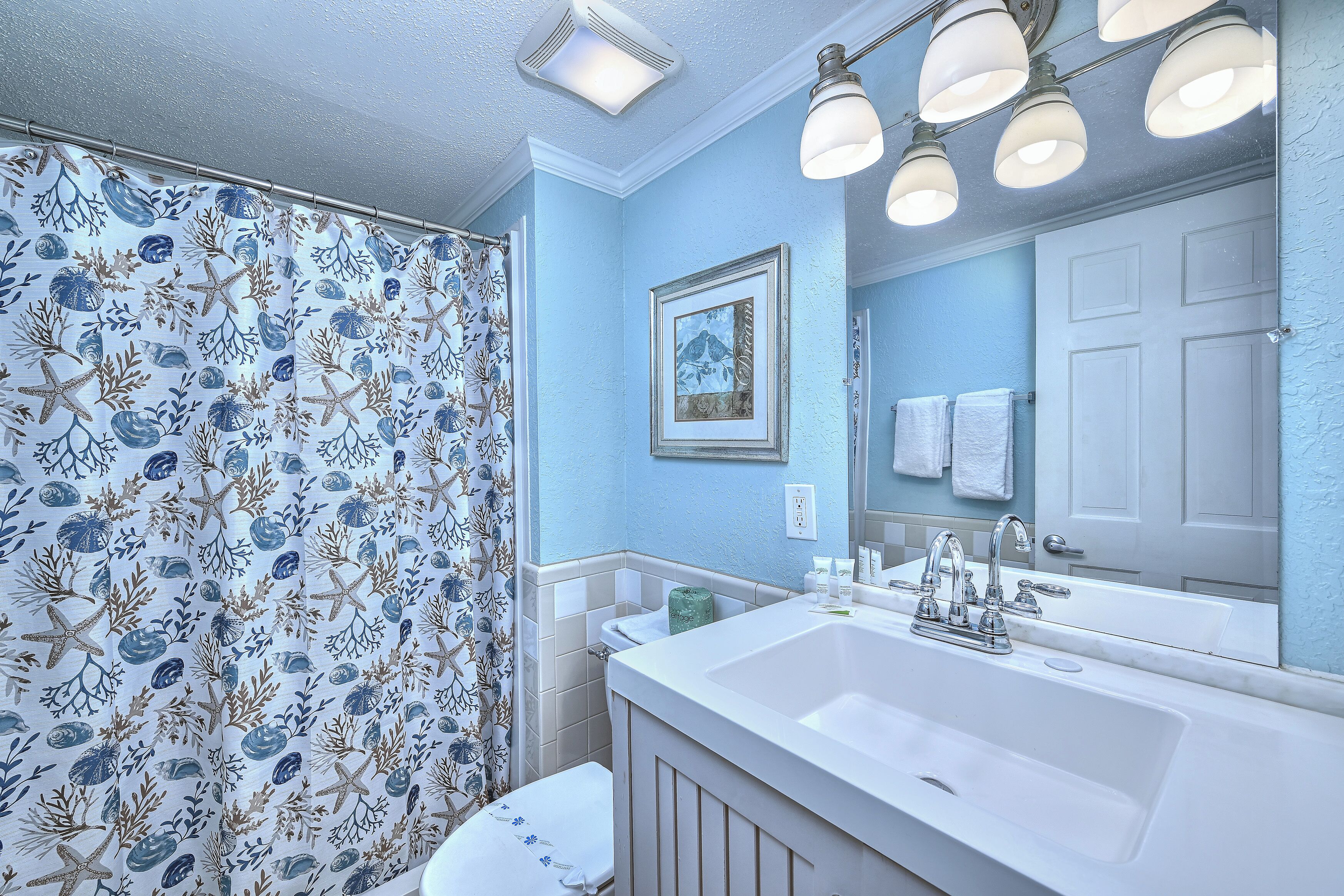 Une salle de bain avec un rideau de douche orné de motifs représentant la vie marine, un lavabo blanc avec un miroir et un tableau encadré accroché au mur.