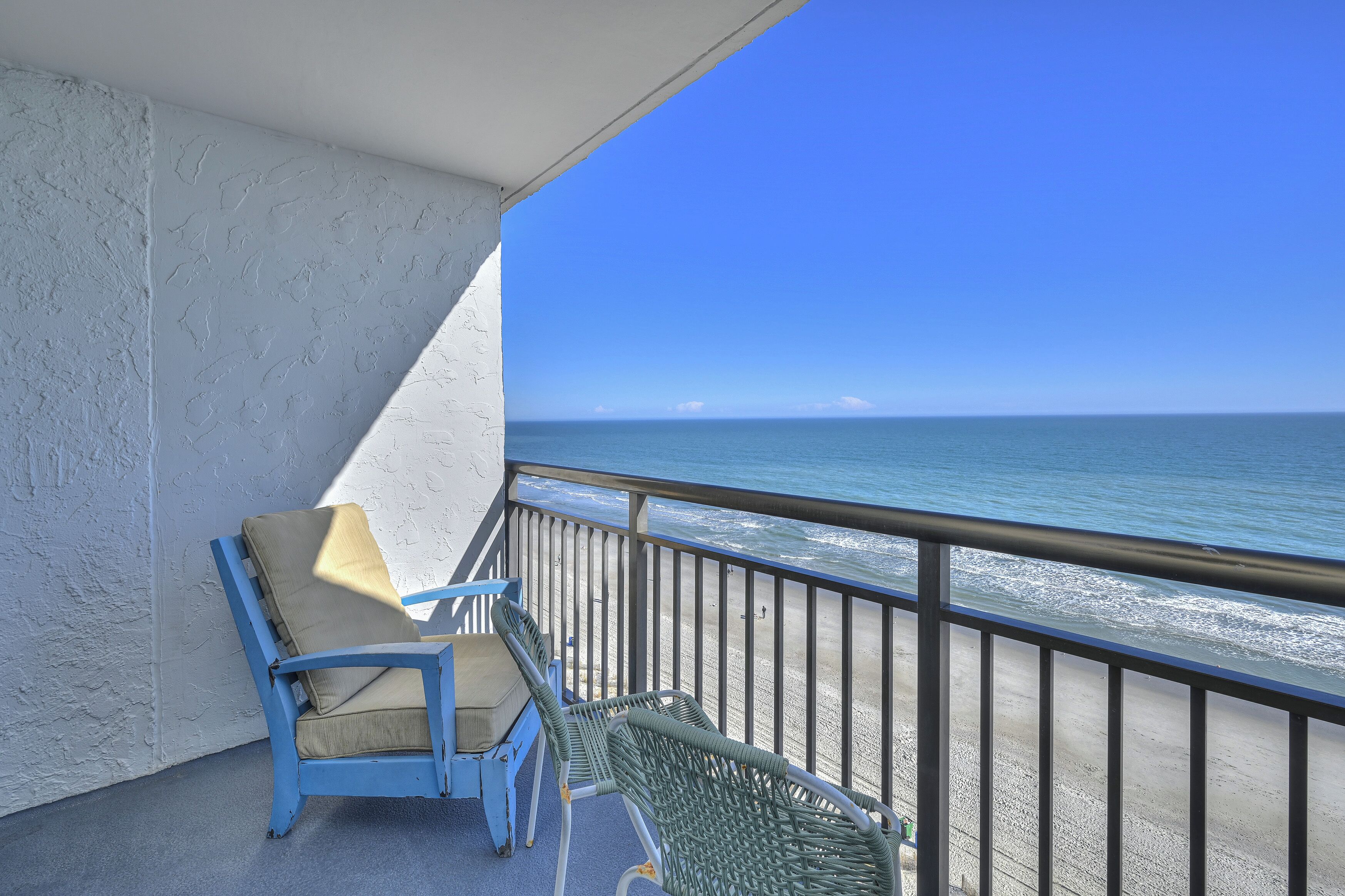 Een balkon met twee stoelen en een tafel, met uitzicht op een strand met een helderblauwe lucht en een rustige zee.