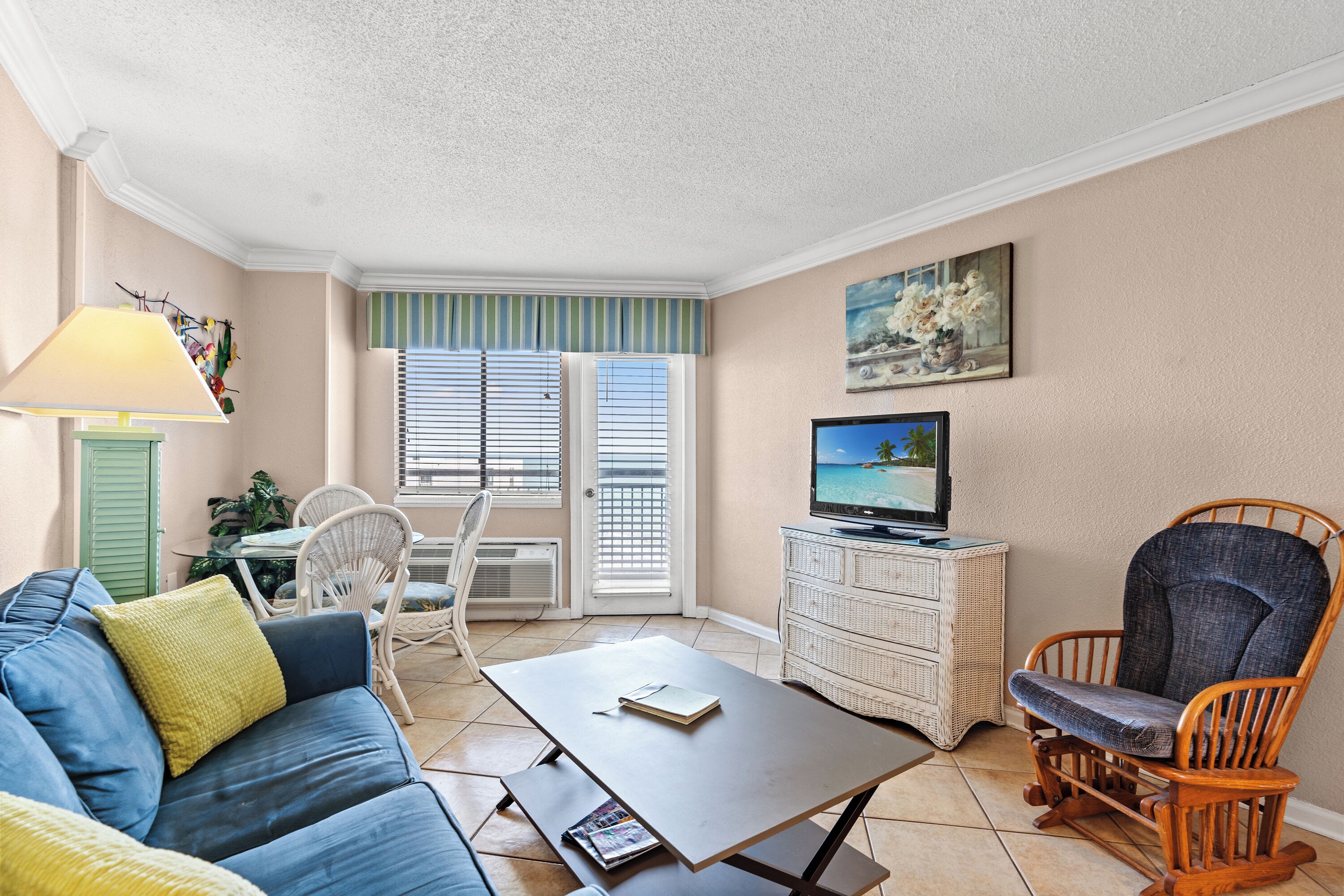 A living room with a sofa, a coffee table, a television, a wicker chair, and a dining area with chairs and a table.