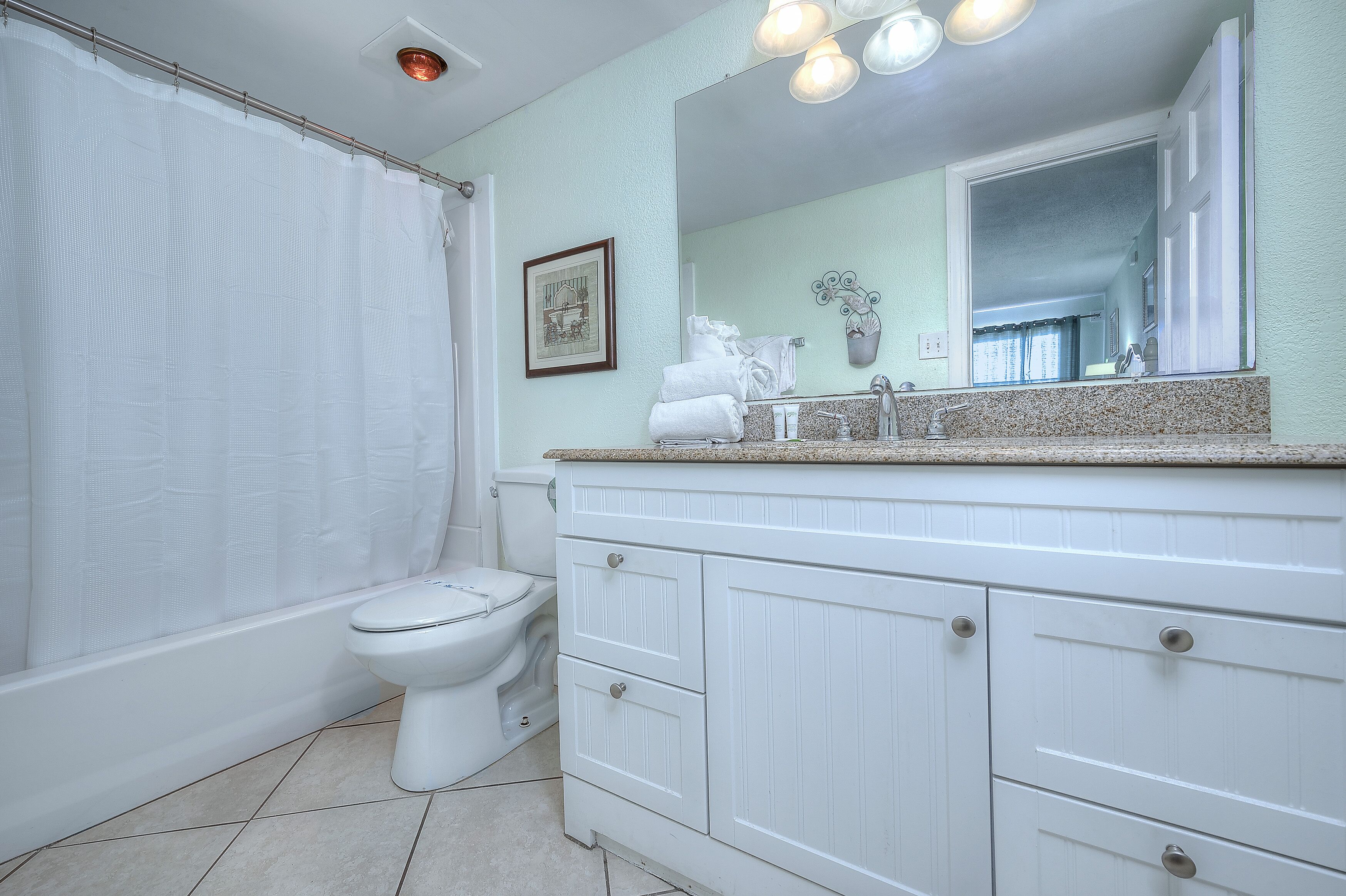 A bathroom with a toilet, a shower curtain, a sink with a mirror, and a cabinet with drawers.