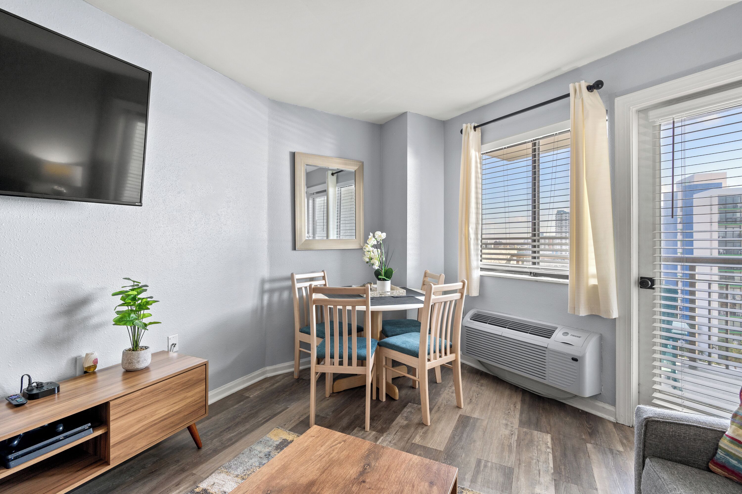A modern living room with a dining table, chairs, a TV mounted on the wall, a potted plant, and a window with blinds.