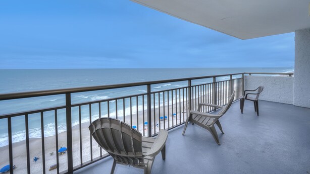 Un balcón con dos sillas Adirondack con vistas a una playa con sombrillas y una vista despejada del océano.