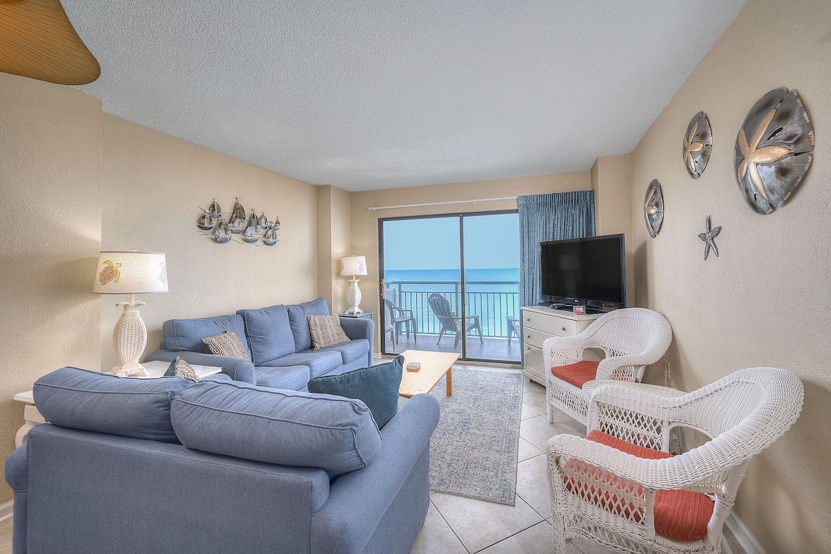 Un salon avec un canapé bleu, des fauteuils en osier blancs, un téléviseur à écran plat et un balcon offrant une vue sur la mer.