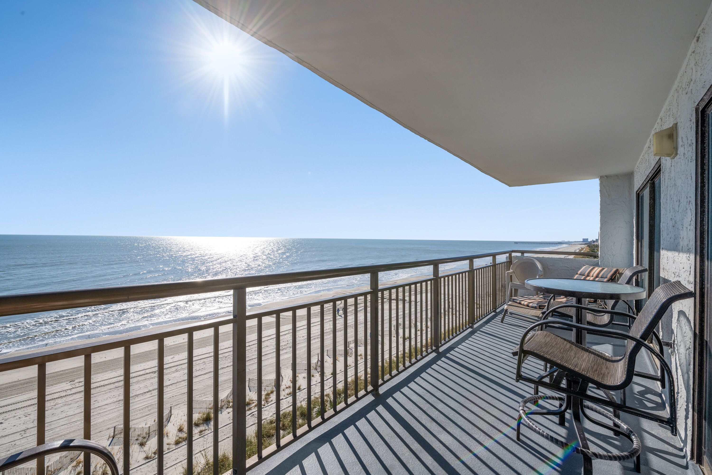 A balcony with a view of the beach and ocean, furnished with a table and chairs.