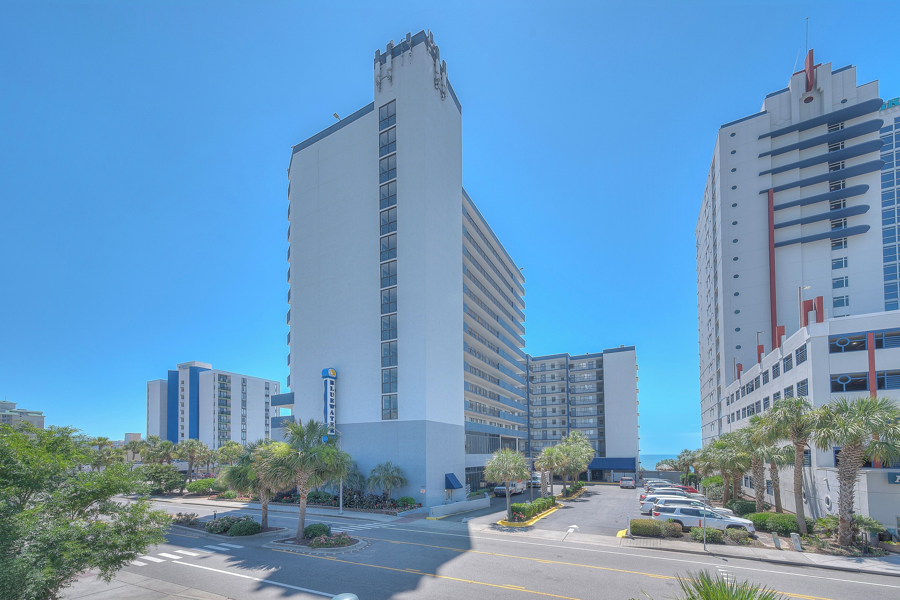 A modern building with a prominent vertical tower, multiple balconies, and a parking lot with cars and palm trees.