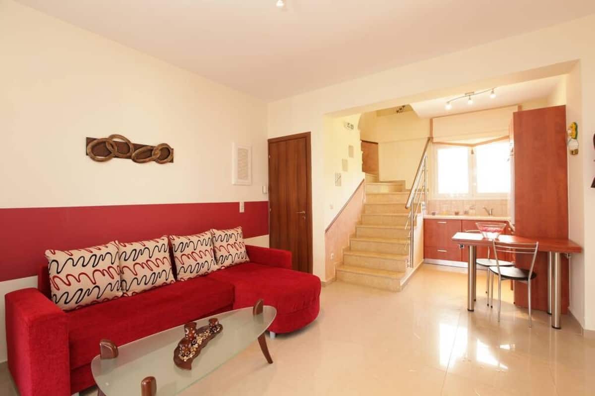 A living room with a red sofa, a glass coffee table, a staircase leading to a kitchen area.