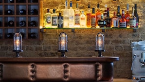 A bar area with a wooden counter, three vintage-style light fixtures, and a variety of liquor bottles displayed on shelves against a stone wall. - The Bull and The Hide (London)