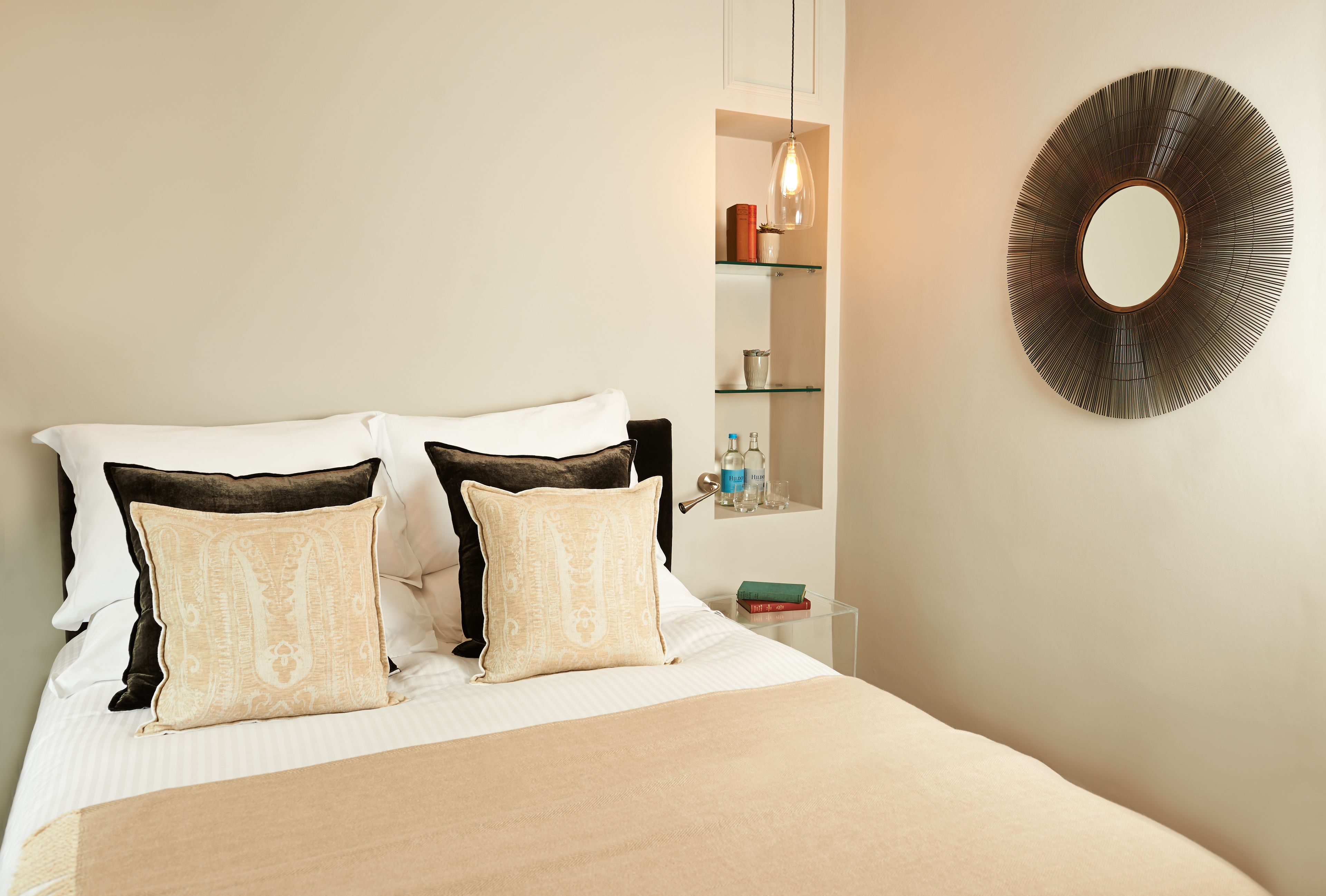 A neatly made bed with two decorative pillows, a bedside table with a lamp, and a wall-mounted mirror.