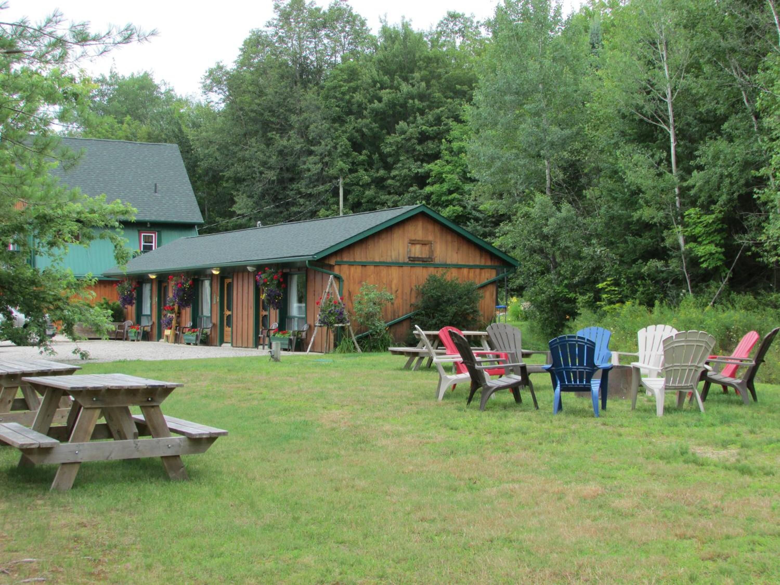 Un área de estar al aire libre rústica con mesas de picnic y sillas Adirondack, una cabaña de madera con techo verde y un denso bosque al fondo.