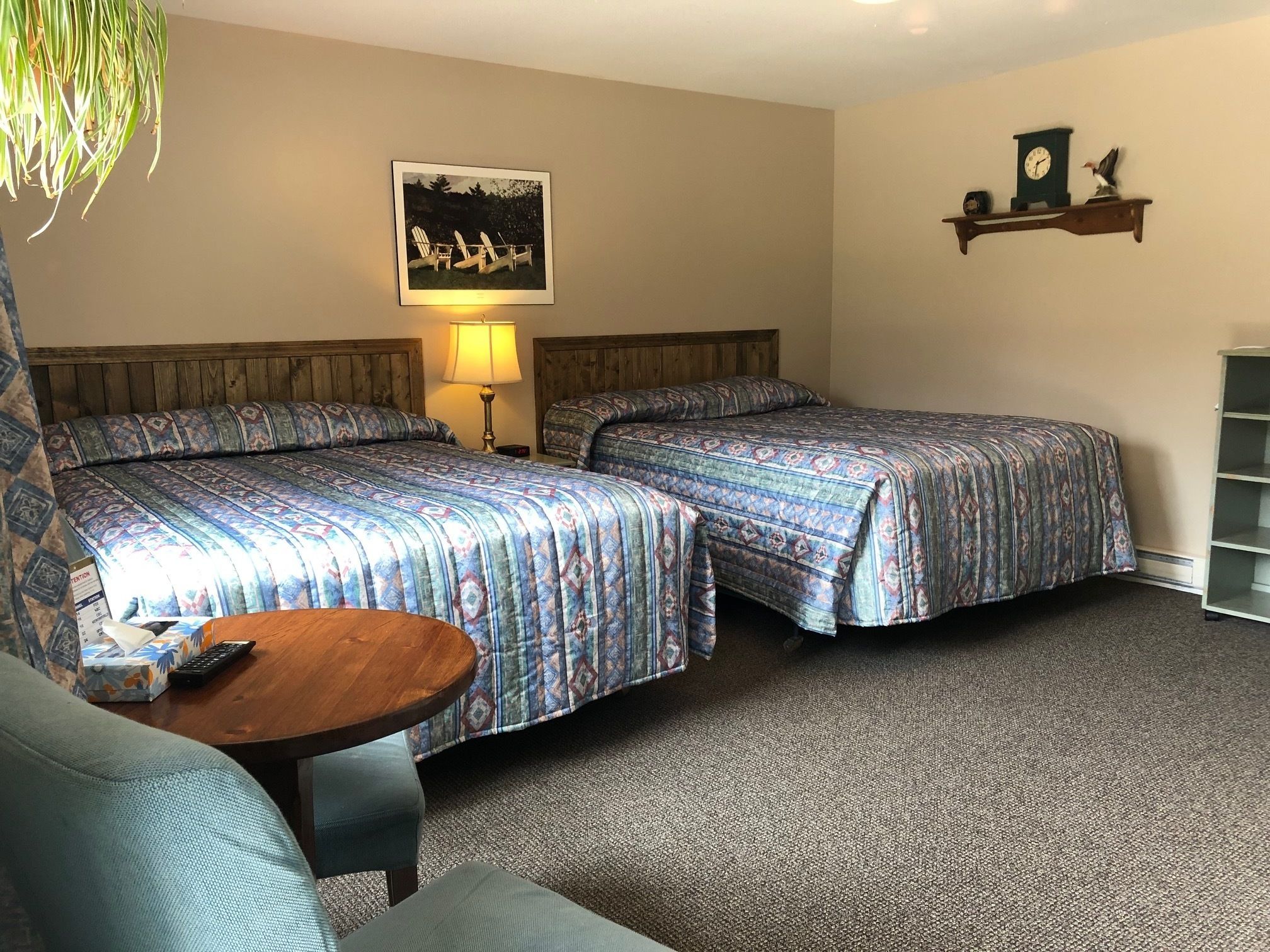 Ein Hotelzimmer mit zwei Betten, einem kleinen Tisch, einem Stuhl, einer Lampe, einer Uhr und einem Regal mit Dekoration.