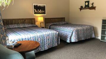 A hotel room with two beds, a small table, a chair, a lamp, a clock, and a shelf with decorative items.