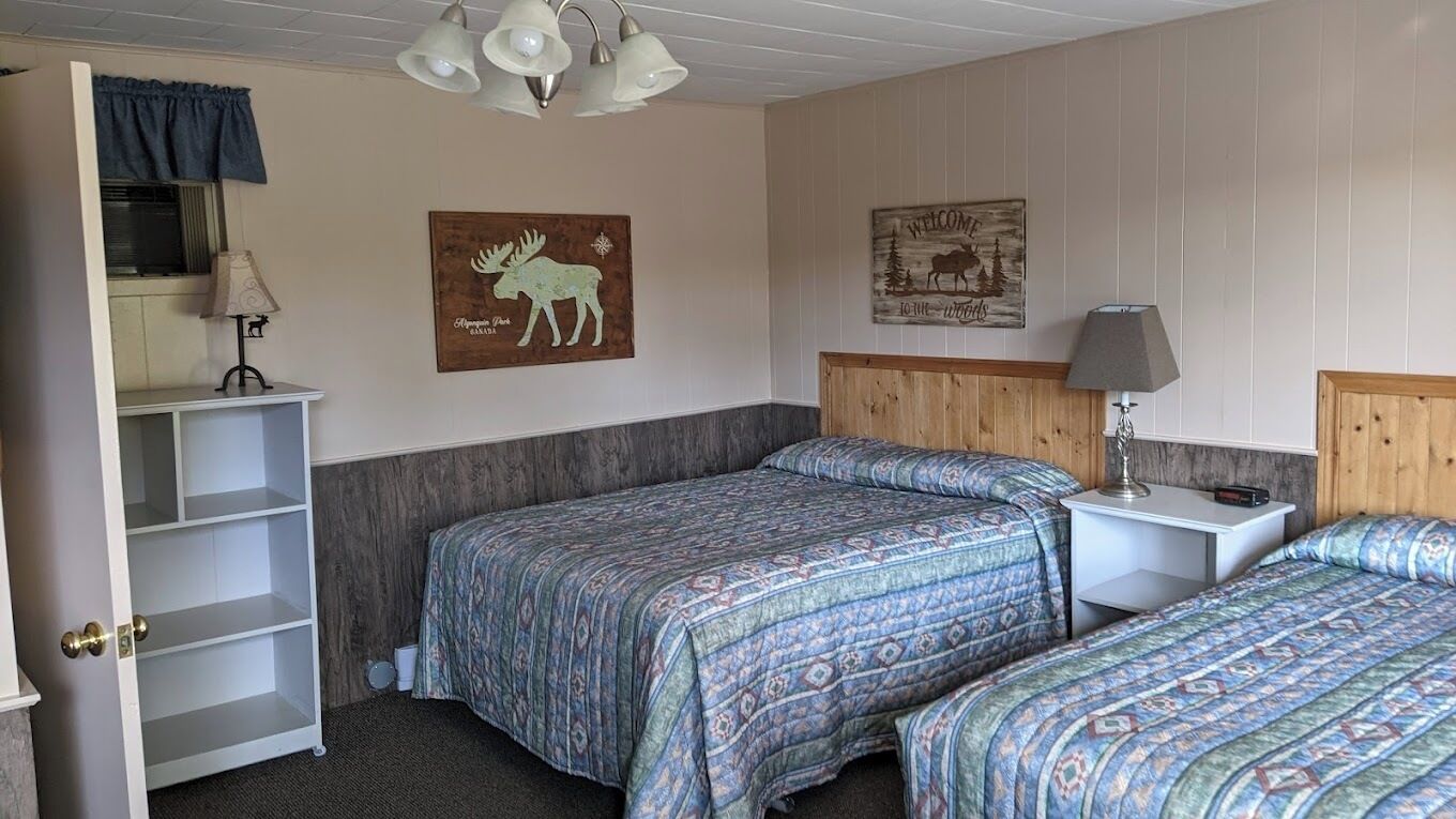 A room with two beds, a wooden headboard, a desk with a lamp, a shelf with a lamp, and two wall decorations.