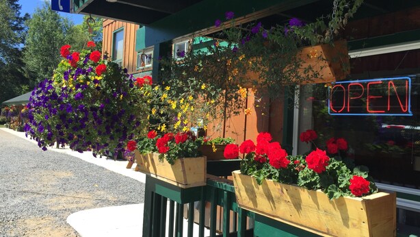 Fachada de madeira, um letreiro de neon com a palavra 'OPEN' e vasos de flores na calçada.