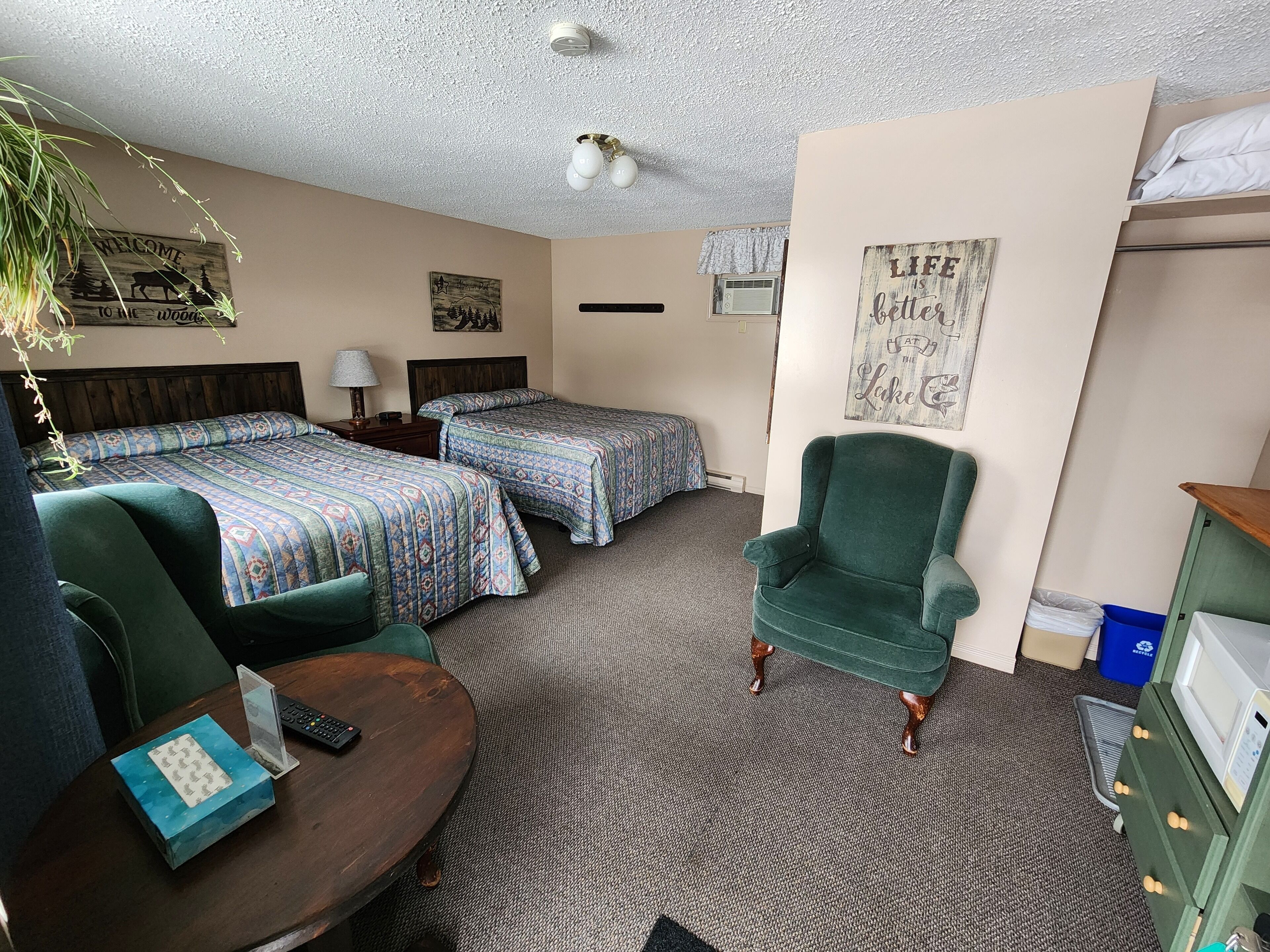Quarto de hotel com duas camas, uma poltrona verde, uma escrivaninha com abajur e uma mesinha de apoio com caixa de lenços.
