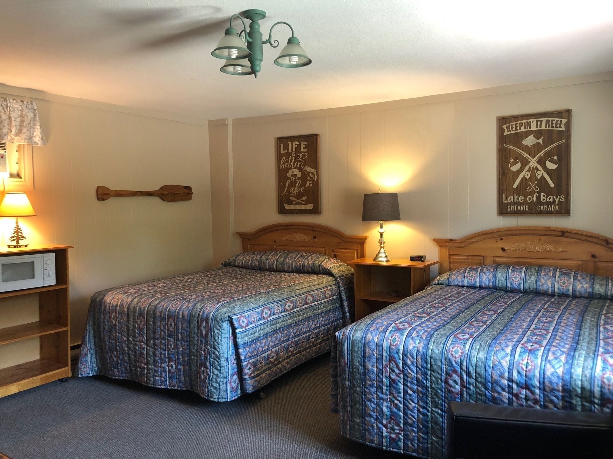 Une chambre d’hôtel avec deux lits, une tête de lit en bois, une lampe fixée au mur, un panneau décoratif encadré et une petite étagère avec une lampe.