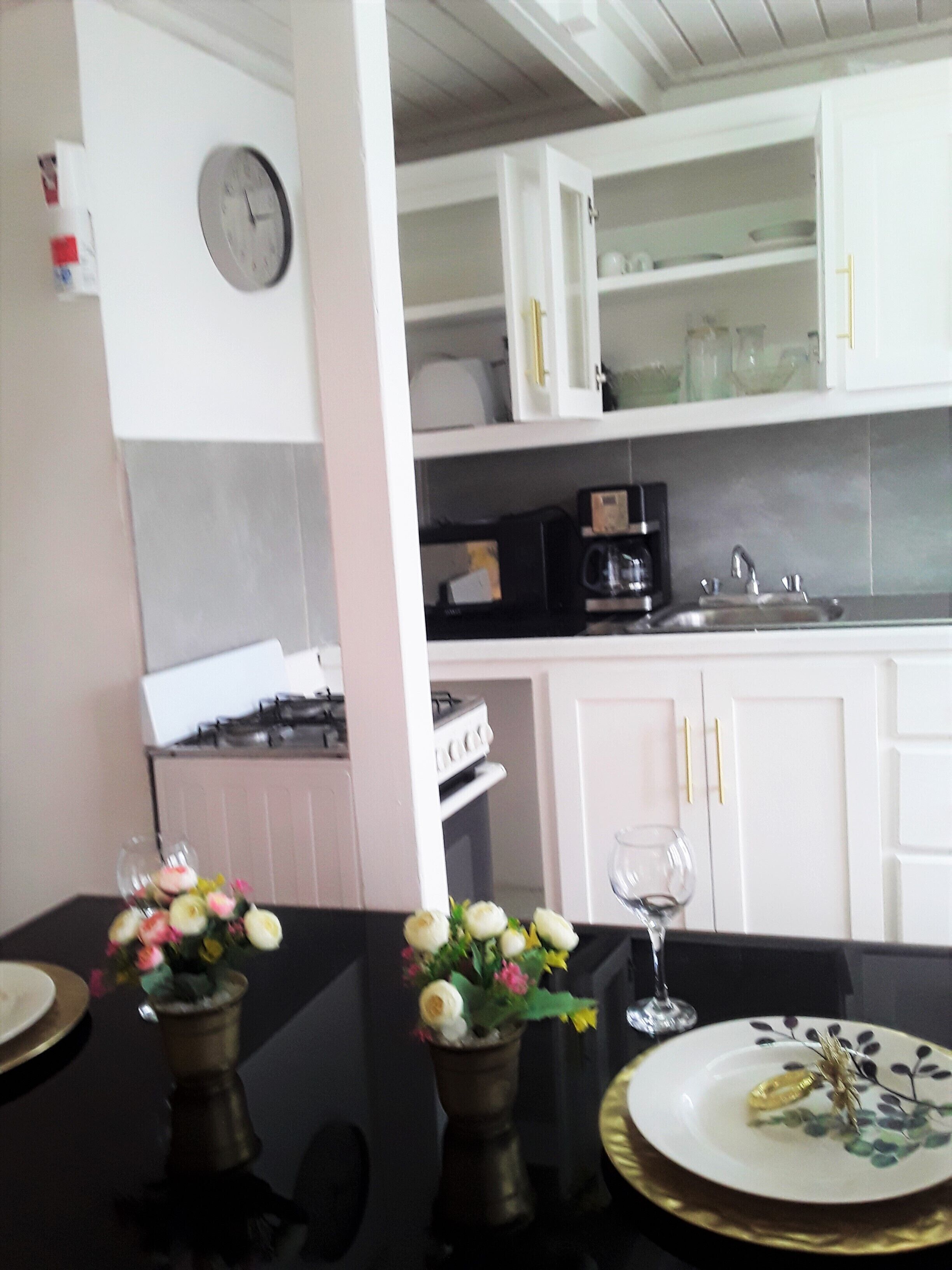 A kitchen with white cabinets, a clock, a coffee maker, and a dining table with a vase of flowers.