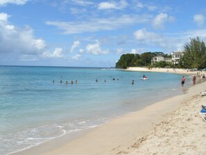 Ein Strand mit klarem türkisblauem Wasser, Menschen beim Schwimmen und Spazierengehen am Ufer sowie eine Reihe von Gebäuden im Hintergrund.