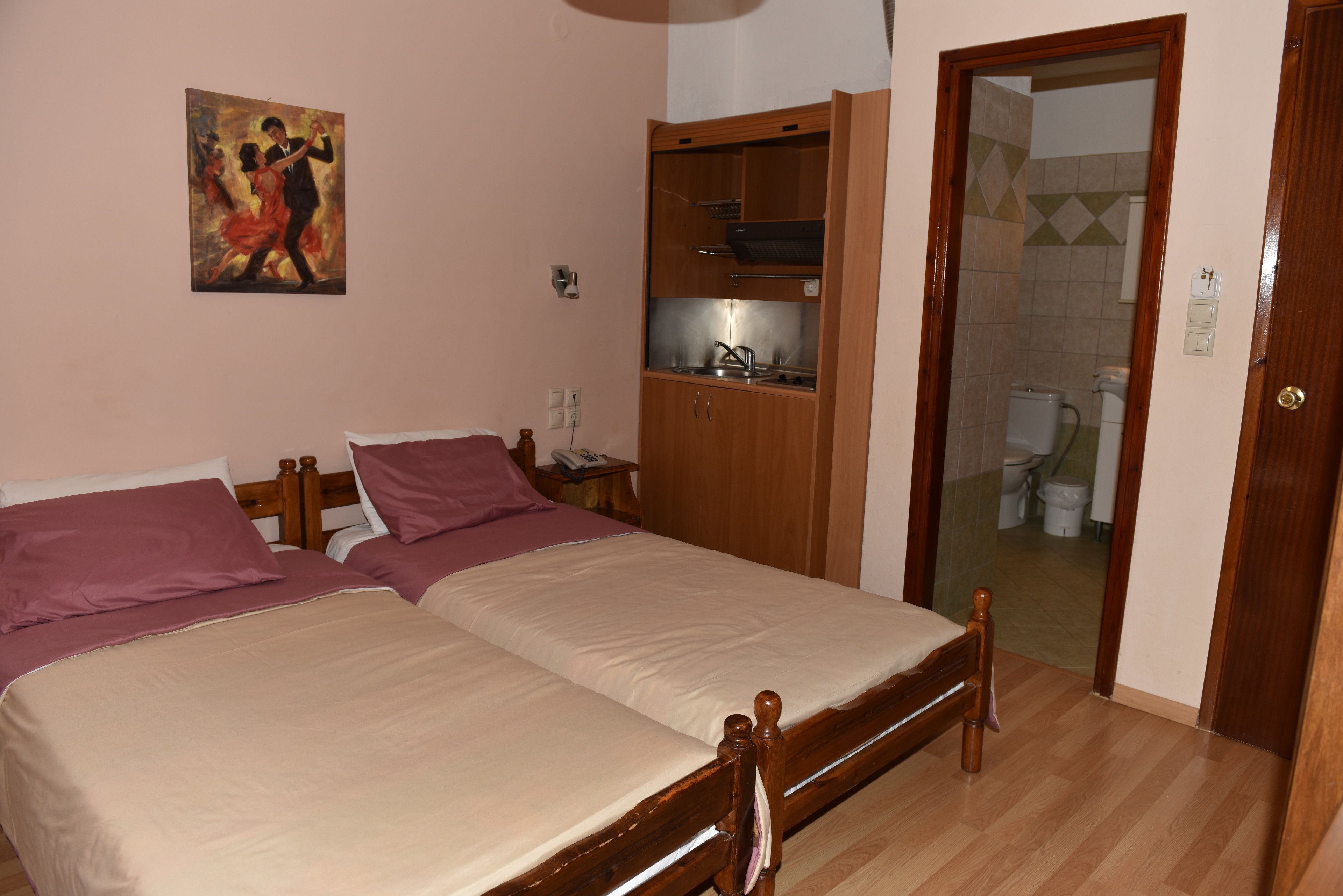 A bedroom with two beds, a wooden wardrobe, a small kitchen area, and a bathroom visible through a door.