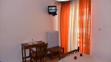 Una habitación con cama, una pequeña mesa de madera con sillas, televisor montado en la pared y una ventana con cortinas naranjas.