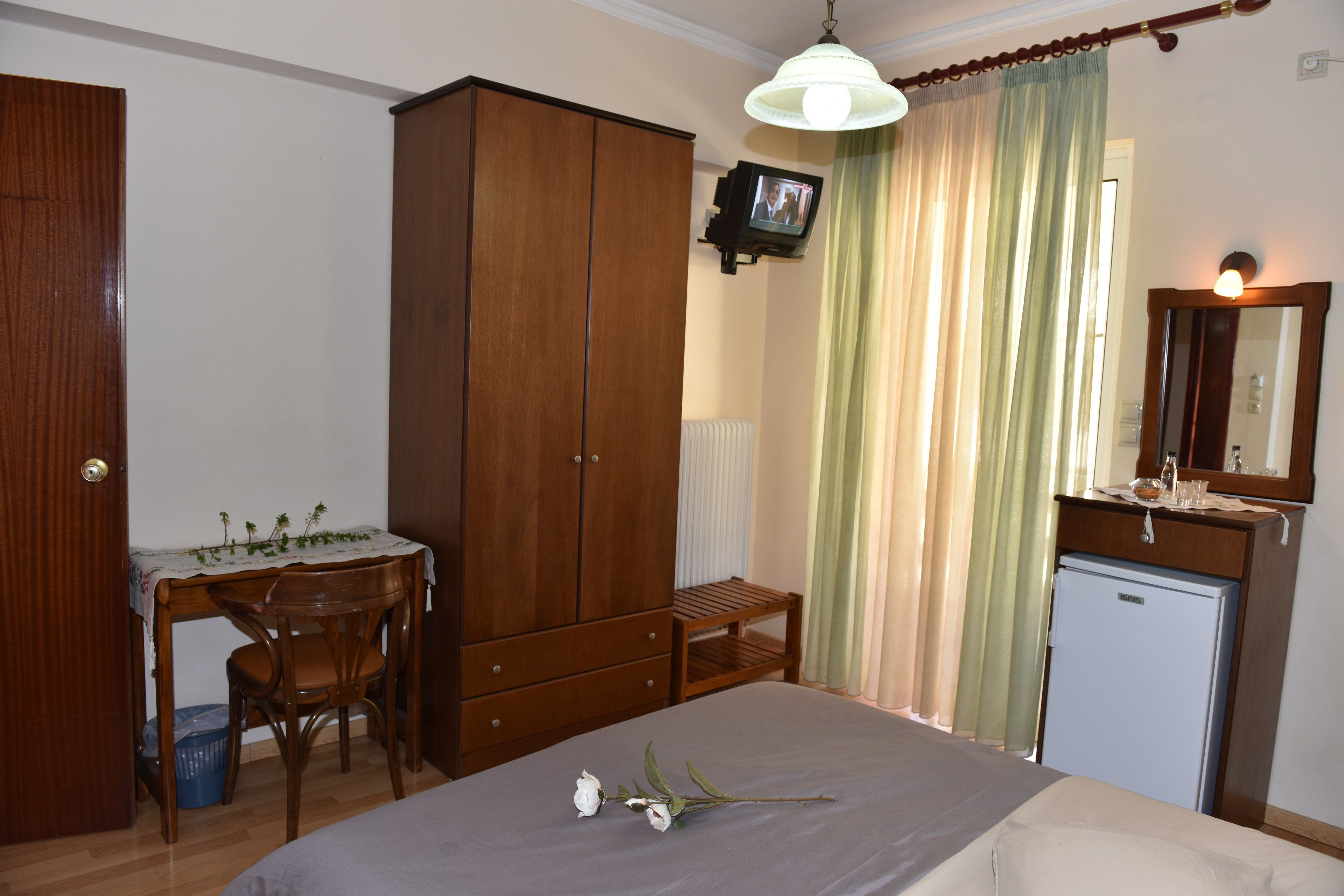 A bedroom with a bed, wooden wardrobe, desk with chair, and a small refrigerator.