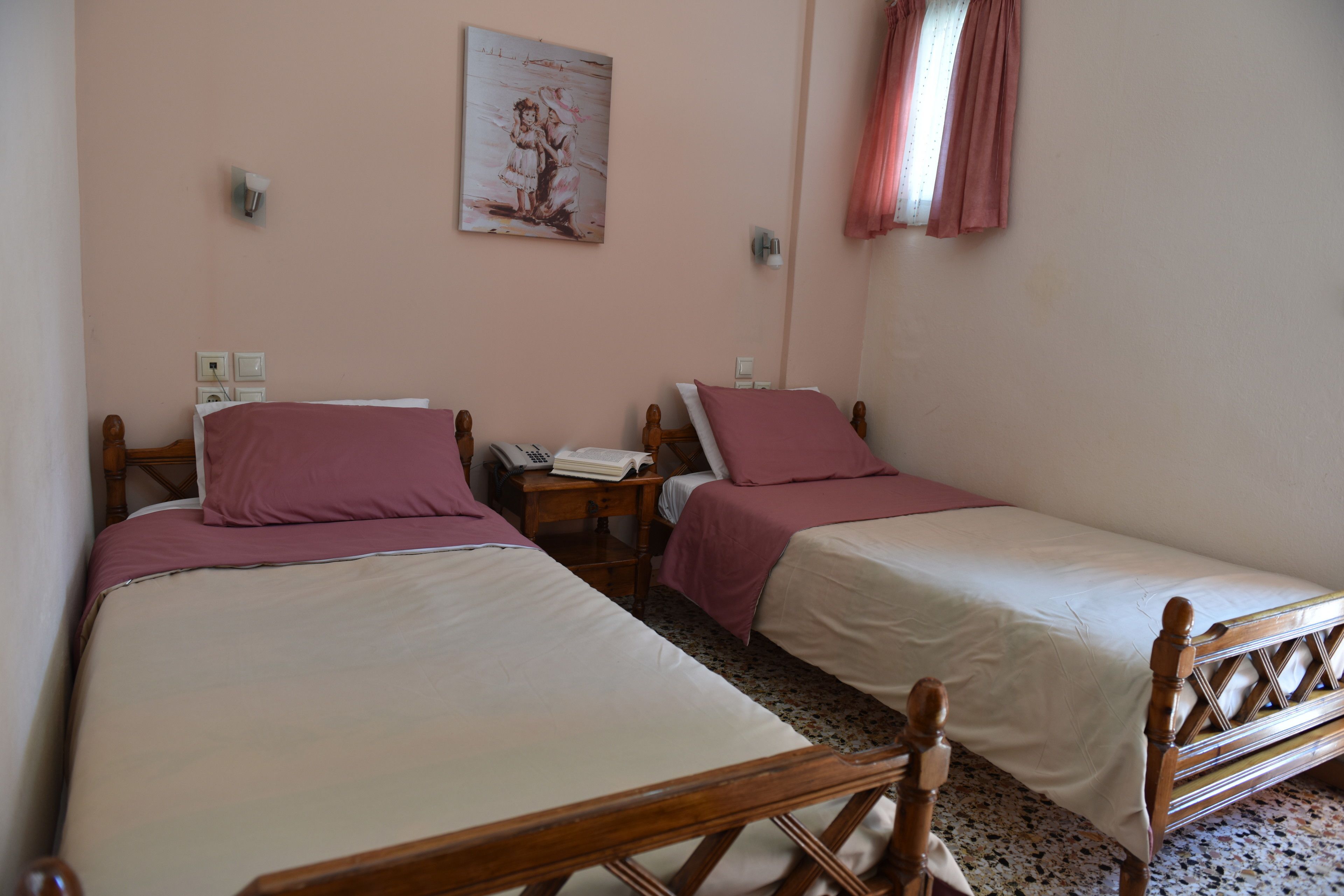 A bedroom with two single beds, each with a wooden frame and pink bedding. There is a nightstand with a telephone and a lamp between the beds. A painting hangs on the wall above the beds.