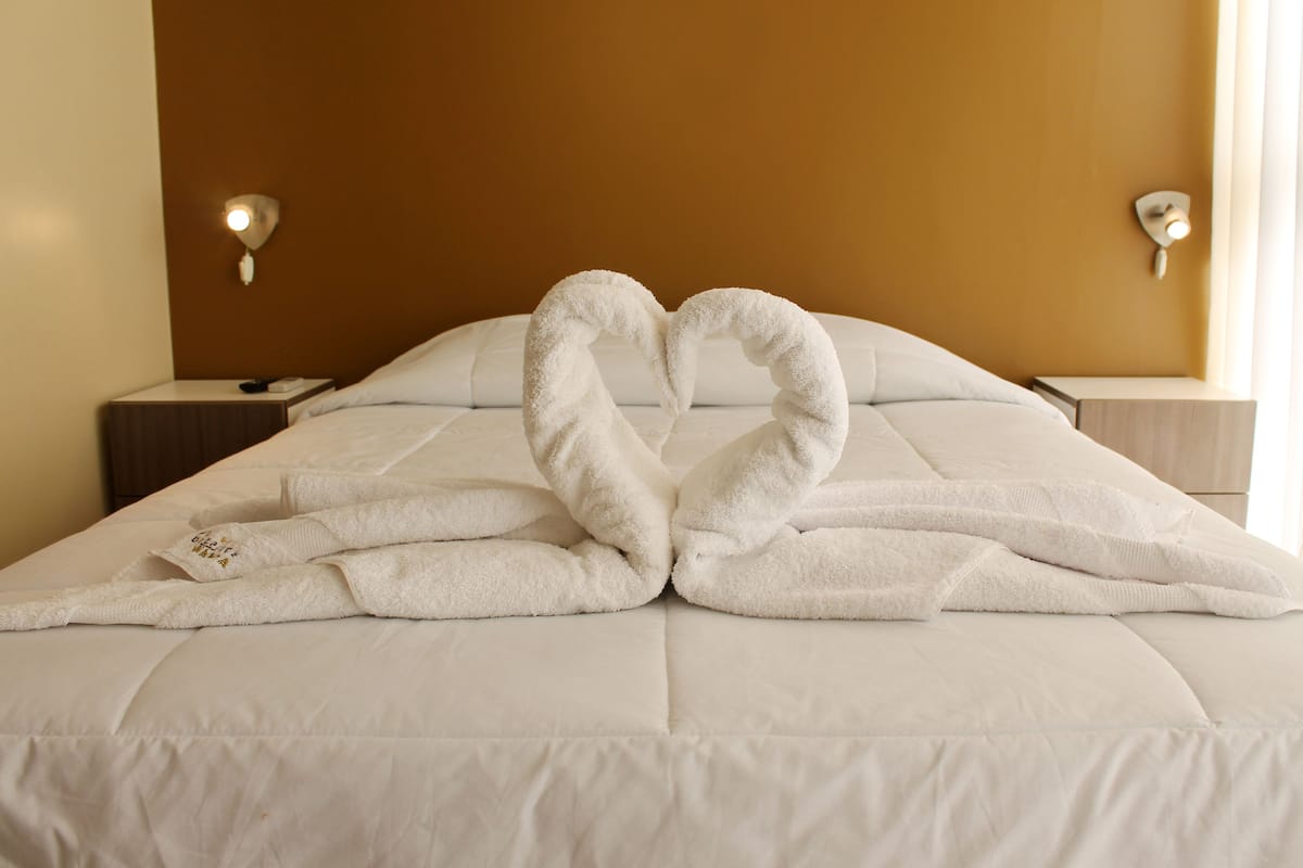 Quarto de hotel com cama, mesa de cabeceira e luminária fixada na parede.