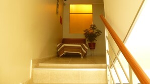 Eine Treppe mit hölzernem Geländer führt zu einer Zwischenebene. Auf der Zwischenebene steht ein Blumentopf, im Hintergrund ist ein Bett mit gemusterten Bettbezügen zu sehen.