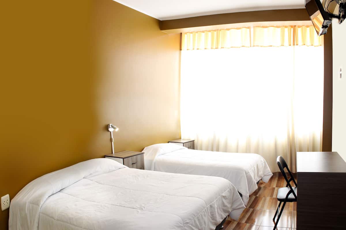 Quarto de hotel com duas camas, uma escrivaninha, uma cadeira, um abajur e uma janela com cortinas.