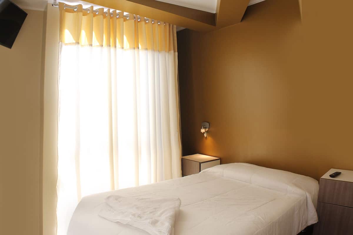 Quarto de hotel com uma cama grande, mesa de cabeceira, luminária e janela com cortinas.