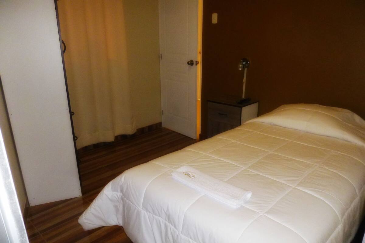 Quarto de hotel com uma cama, mesa de cabeceira, abajur e uma porta que dá para outro espaço.