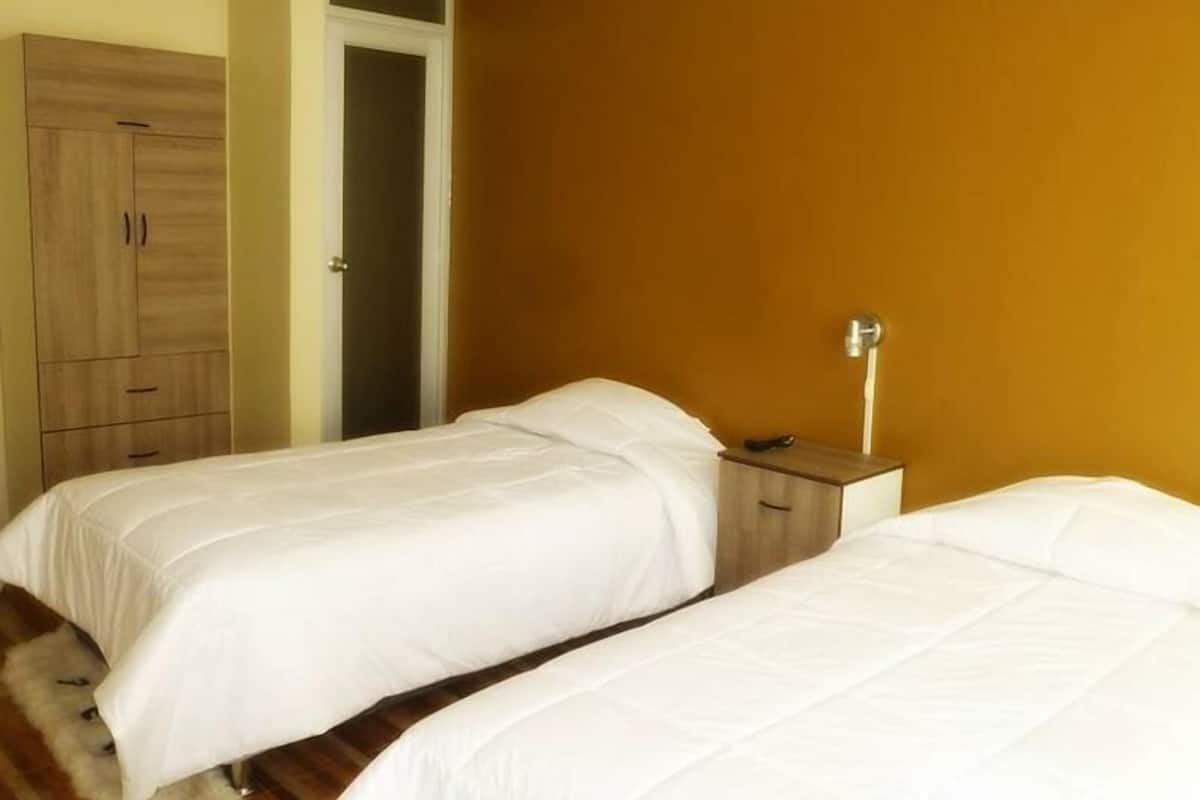 Quarto de hotel com duas camas de solteiro, um guarda-roupa de madeira e uma mesa de cabeceira com abajur.
