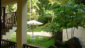 A view from a balcony with a wooden railing, overlooking a garden area with a white umbrella table and chairs, surrounded by greenery and rocks. - Pousada 3 Bacias (Resende)